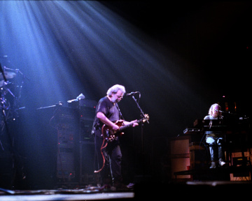 Jerry Garcia soloing onstage with the Grateful Dead. vince Welnick is in the background, and Jerry is illuminated from above by a spotlight